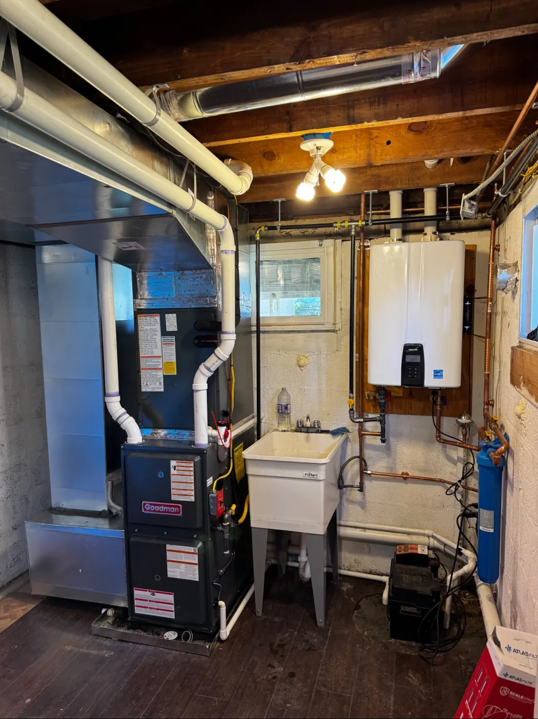 A basement with a Goodman furnace, white PVC venting and a tankless water heater mounted on the wall beside a utility sink