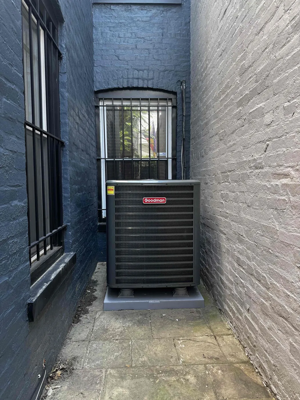 A Goodman condenser on a new pad in a narrow brick alley beside a rowhouse