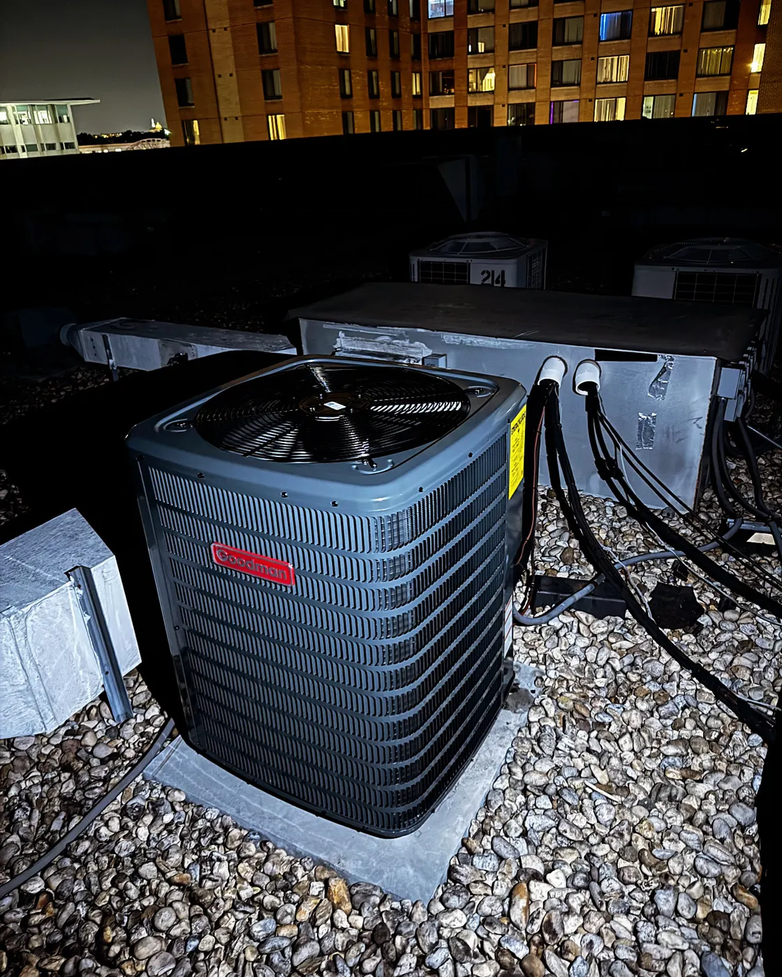 A new Goodman condenser on a gravel rooftop at night with city buildings lit up behind it
