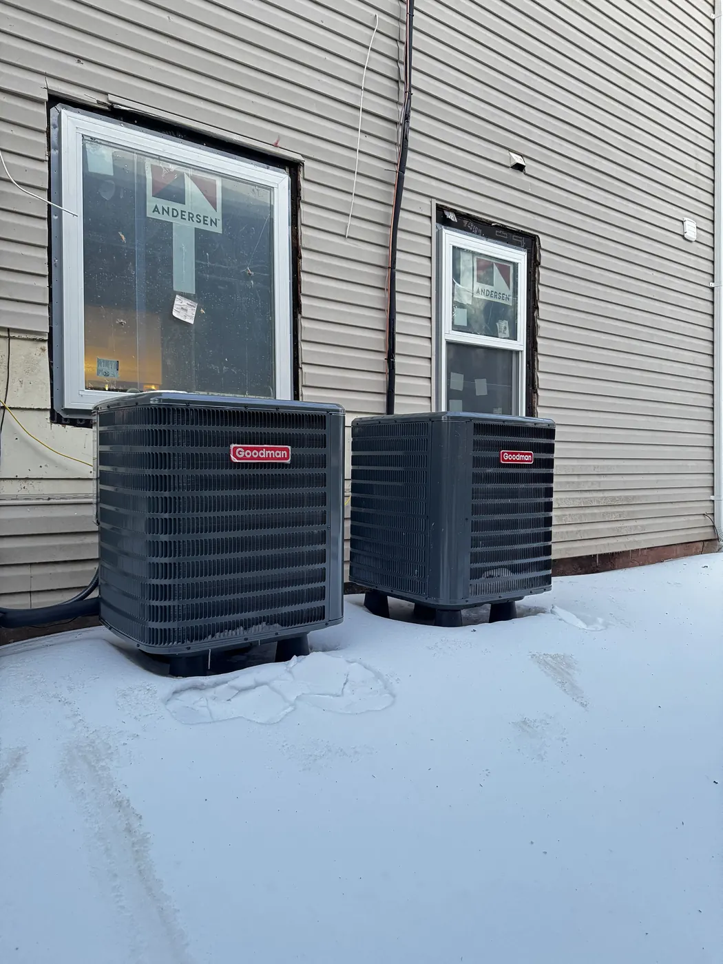 Two new Goodman condensers side by side on a white flat roof beside gray siding