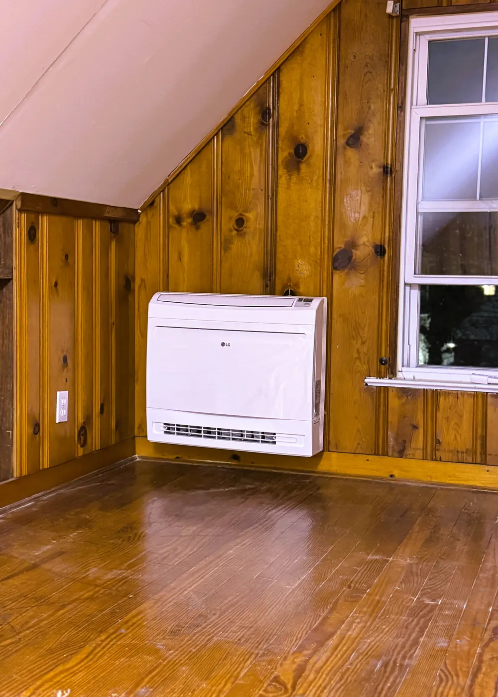 A white floor-level ductless unit installed under a window in a knotty pine room