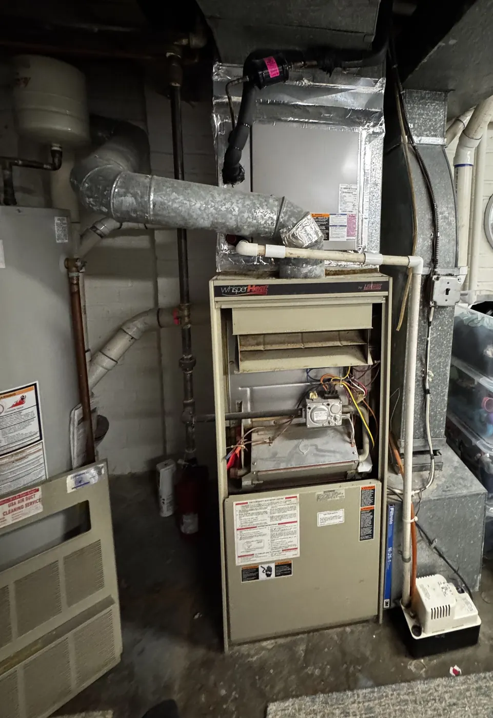 An older gas furnace with its panel off in an unfinished space, flex duct and venting above it