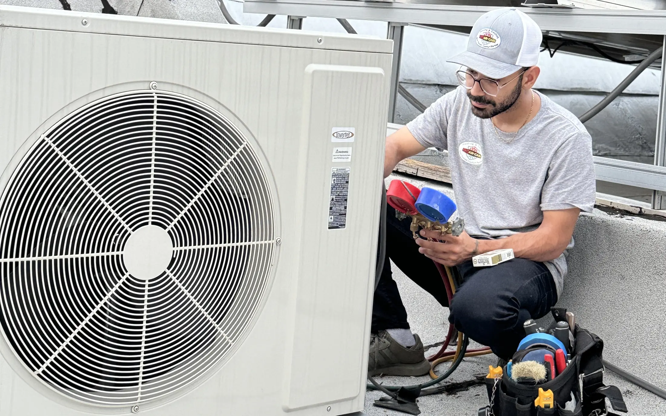 A 911Air-Care tech in a company shirt and cap kneeling on a flat roof, reading red and blue refrigerant gauges at a ductless outdoor unit