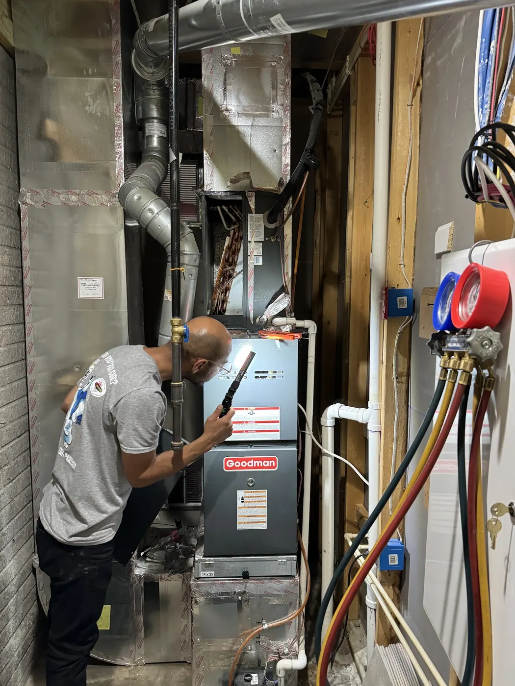 A 911Air-Care tech in a company shirt checking a Goodman furnace in a tight utility closet with a flashlight